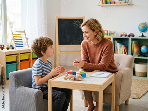 Child psychologist or female teacher talking with little boy during therapy session in cozy classroom, early education concept
