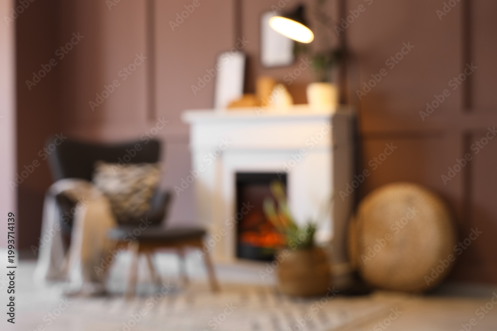 Naklejka premium Blurred view of cozy living room with grey armchair, footstool, glowing lamp and stylish fireplace