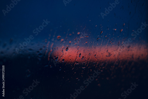 Rain on glass during sunset creates a moody blue and red scene, with droplets in the foreground and a distant horizon under twilight skies as night approaches