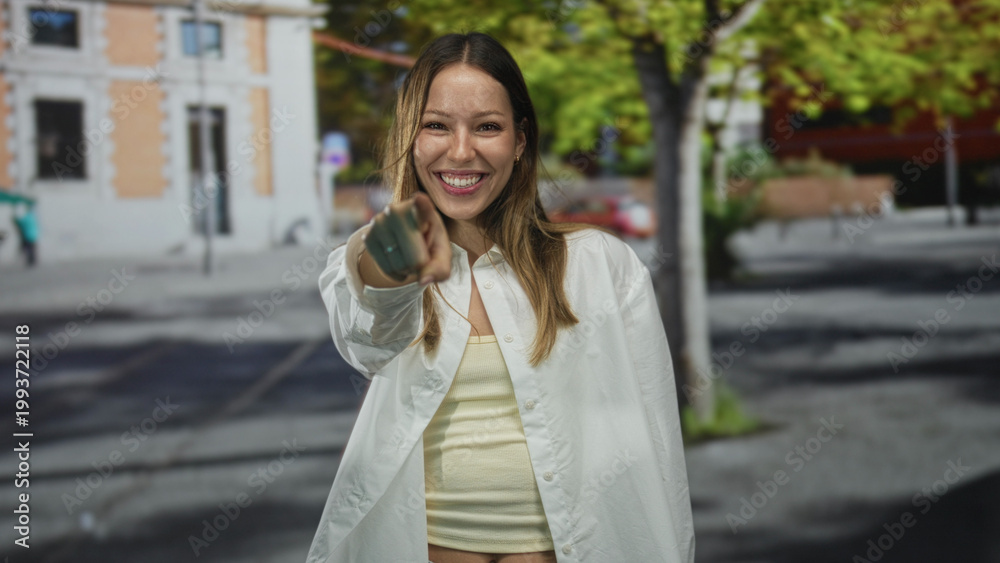Fototapeta premium Young woman pointing finger at camera and smiling on street with building and trees in frame; playful invitation.