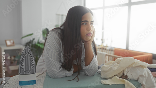 Woman ironing clothes on ironing board with steam iron and pile of laundry inside a home building nursery, leaning on board with tired expression; quiet fatigue.