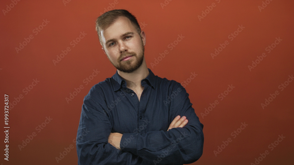 Obraz premium Young caucasian man with arms crossed in a studio against a burnt orange backdrop wearing a navy button shirt and light beard; confidence.