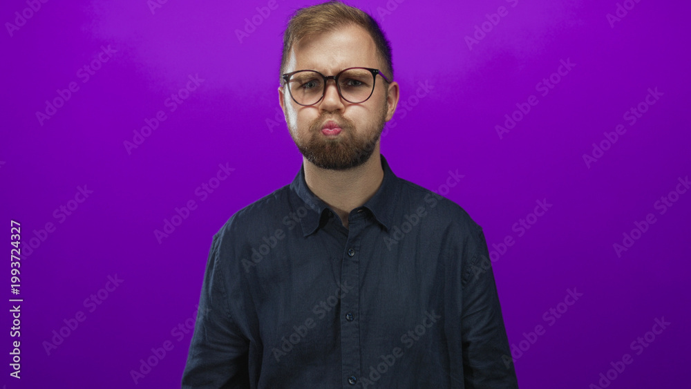 Obraz premium Man with glasses puffing cheeks in studio against a purple backdrop, young bearded and wearing dark shirt; playful moment.