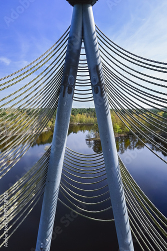Cable bridge structure over a calm river (at Lommel - Belgium), in which trees are reflected under a blue sky.