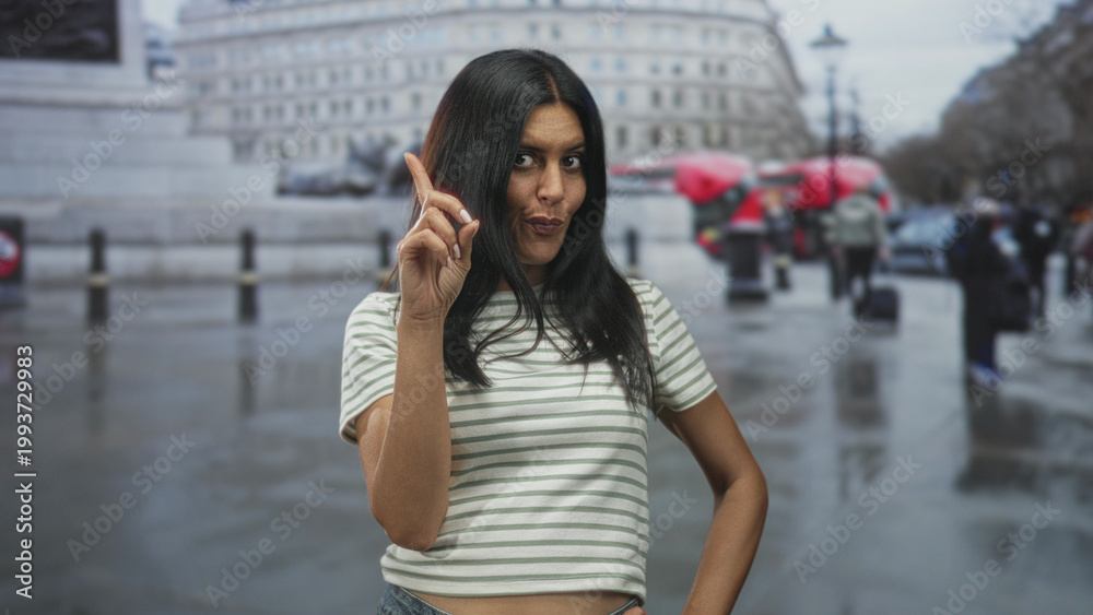 Fototapeta premium Woman points finger with hand on hip, midriff visible and slight smirk while standing on a wet city street near red buses and bollards; confidence.