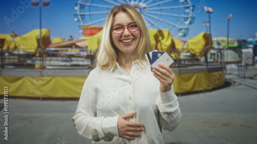 Photos Woman holding creditcards, hand displaying cards and other hand on stomach in street fair with ferris wheel and bright tents; shopping joy