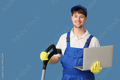 Young male janitor with modern vacuum cleaner and laptop on blue background