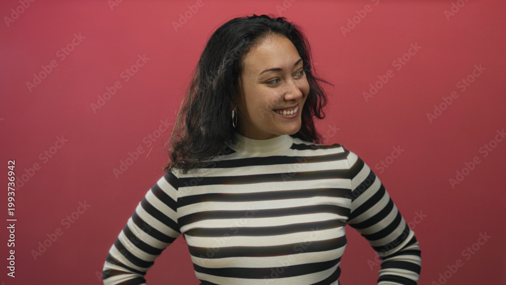 Fototapeta premium Young hispanic woman smiling with hands on hips in red studio wearing black and white striped turtleneck and hoop earrings; confidence.