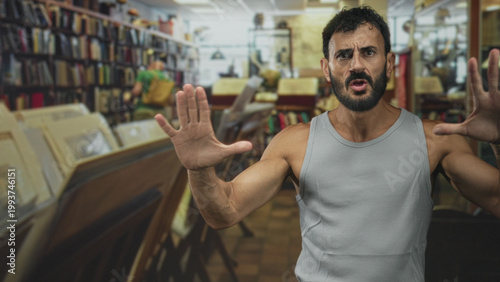 Tableau sur toile Man with hands raised and visible forearms, tense face, standing among bookshelves and framed prints inside a bookstore building; alarm defiance