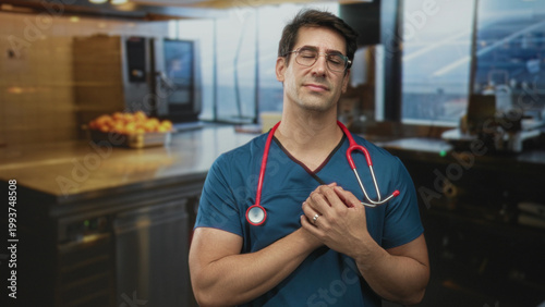 Photos Doctor man in blue scrubs with red stethoscope holds hands over heart in airport lounge kitchen near counter and fruit tray; compassion fatigue