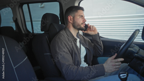 Man holding smartphone to ear while seated at steering wheel inside a car beside an industrial building, talking and gesturing with his free hand; concern.