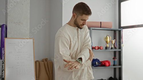 Cuadro en lienzo Man adjusting wristwatch and stretching forearm while rubbing elbow near shelves with boxing gloves and trophies in a training studio; focus preparation