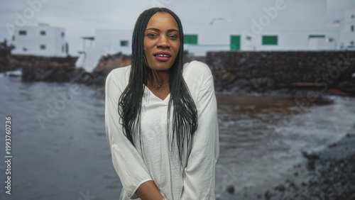 Billede på lærred Woman in white blouse hugs herself at a seaside building, braided hair and smartwatch visible; pensive solitude