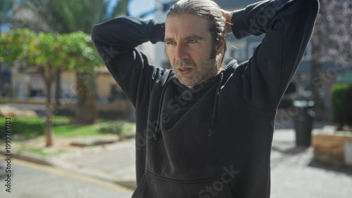 Canvas Print Man with hands behind head touching hair and briefly holding smartphone on street; concern contemplation decision
