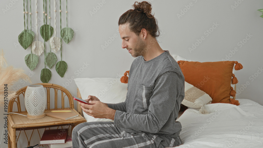 Fototapeta premium Man sitting on bed in gray loungewear reaching with right hand for red smartphone on wicker nightstand beside pillows and macrame wall hanging in studio; calm morning.