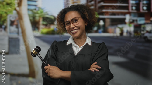 Fotografía Woman judge holding gavel with crossed arms on street smiling, wearing robe and glasses; justice confidence