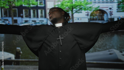 Canvas Print Man priest wearing clerical collar and cross necklace, arms outstretched on a street by a canal; devotion reflection serenity