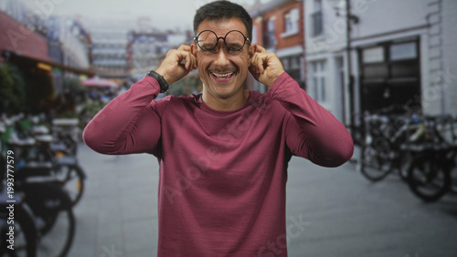 Quadro em tela Young hispanic man with moustache and round glasses tugs his ears with fingers, smiling in a city street with parked bicycles and storefronts; playful joy