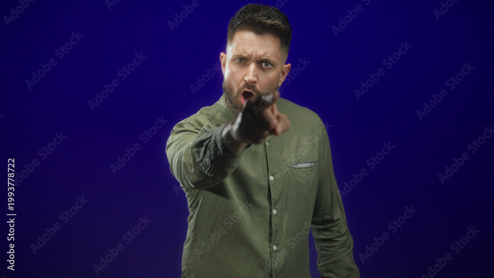 Fototapeta premium Hispanic man with beard thrusts clenched fist toward camera in studio with olive shirt, stern gaze and scowl; anger confrontation.