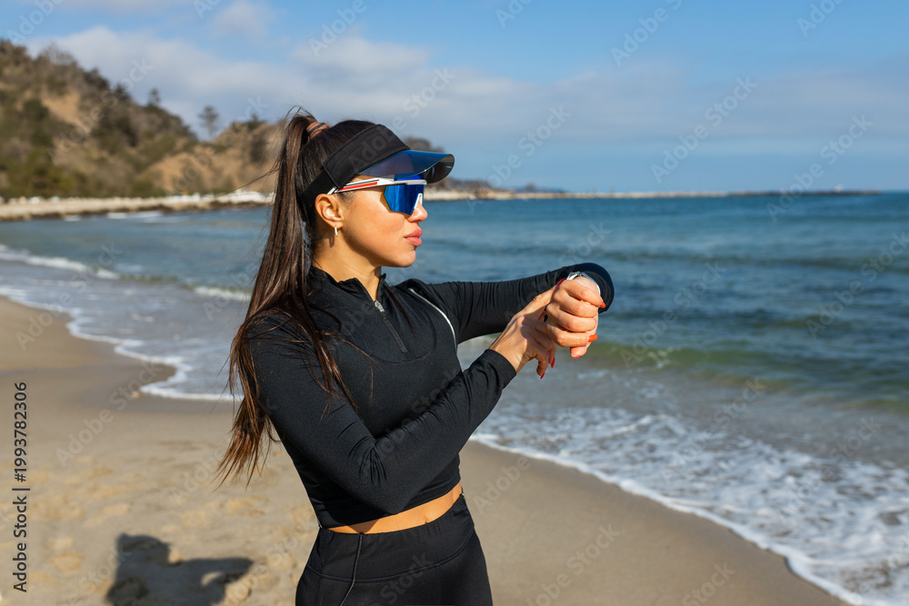 Obraz premium Fit woman checking smartwatch on beach, active lifestyle, outdoor workout by sea, fitness tracking concept