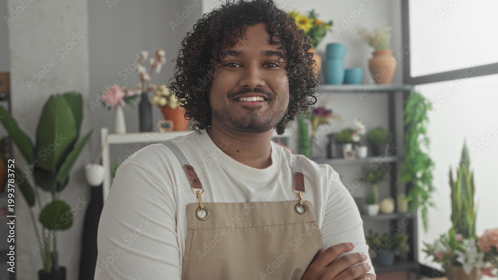 Fototapeta premium Man florist wearing apron holding flowers, arms crossed and smiling near potted plants and vases by a bright window in a shop building; pride craft.