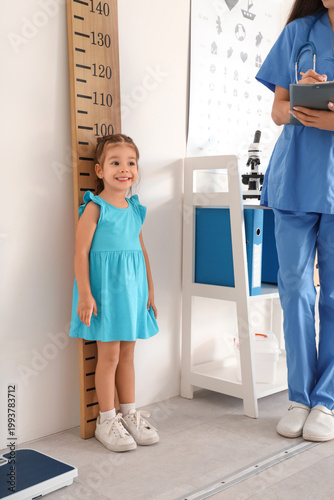 Female pediatrician with cute little girl measuring height near stadiometer i...