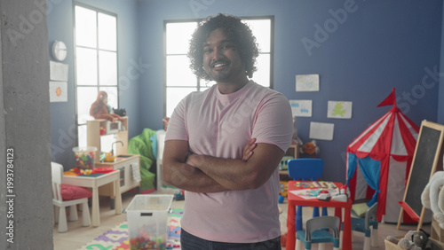 Man with arms crossed smiling in a kindergarten playroom building with red play tent and scattered toys; warmth care learning.