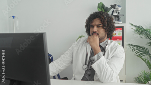 Принти на полотні Man doctor with stethoscope at computer, studying screen, rests hand on chin in a building; thoughtful concern