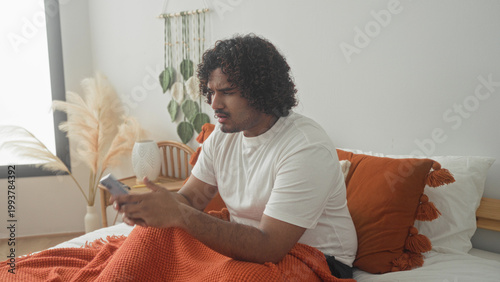 Fotografie Man sitting on bed covering face with hands, orange blanket and pillow and bedside vase visible inside building; fatigue introspection