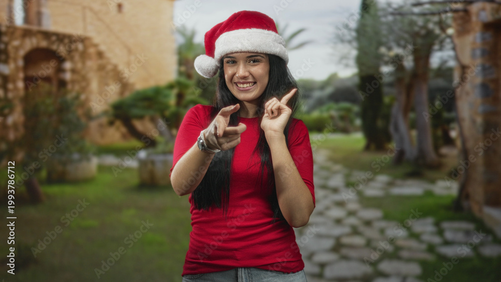 Fototapeta premium Woman pointing finger to camera with hand and smiling while wearing santa hat and red shirt in a historic stone courtyard with arched building and path; holiday joy.