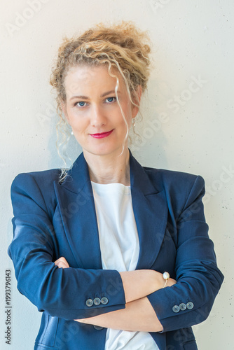 Business woman in blue blazer and white shirt leaning against wall with arms crossed, smiling confidently, formal corporate portrait with copy space