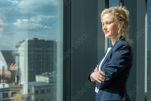 Business woman standing with arms crossed by large window with city view, blue blazer and white shirt, confident corporate lifestyle with copy space