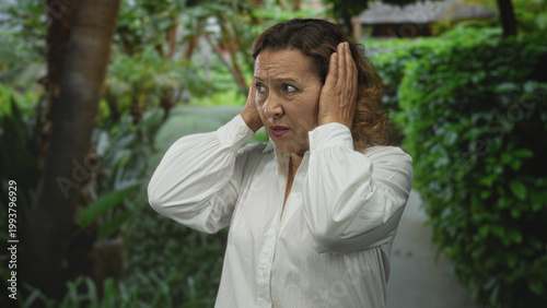 Foto Woman covers her ears with both hands in a lush green outdoor park pathway surrounded by dense foliage; noise discomfort annoyance