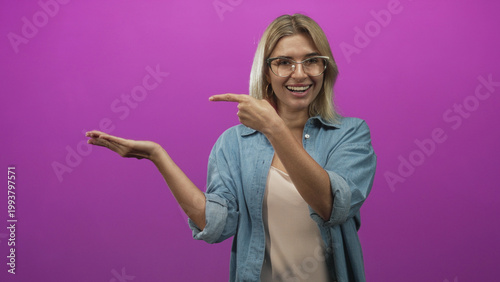 Photos Woman presenting palm and pointing with her other hand in studio purple backdrop wearing denim shirt and glasses; friendly joy