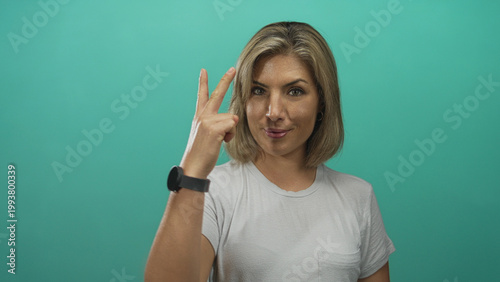 Obraz na płótnie Young blonde woman displays victory sign with raised hand in turquoise studio; triumph confidence spirit