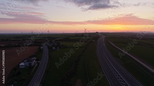 Aerial drone footage at sunrise showing the A30 dual carriageway cutting through the countryside, with wind turbines on the horizon beneath a vibrant, colourful sky