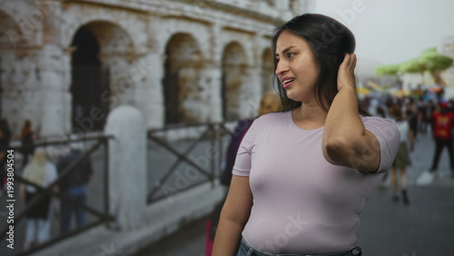 Fotografía Woman with hand to hair and windblown hair, wearing short sleeve tshirt near the roman coliseum building among tourists on a crowded street; travel annoyance