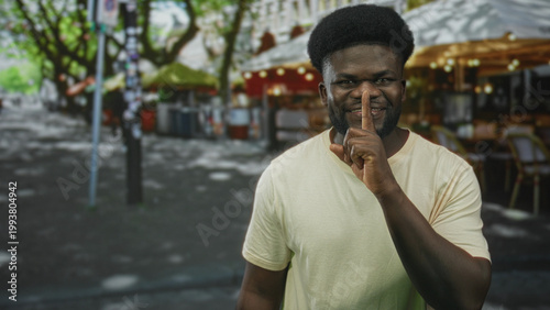 Fotografie Young man smiling points finger and finger to lips for silence while standing near outdoor cafe on a city street; playful secrecy