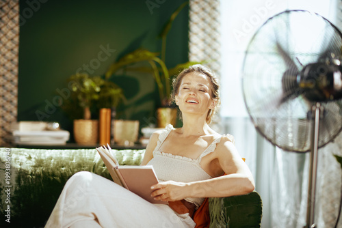 Konstfotografi A joyful woman, eyes closed and smiling, finds serene comfort reading a book on a green velvet sofa, enjoying the sun and a cooling breeze indoors