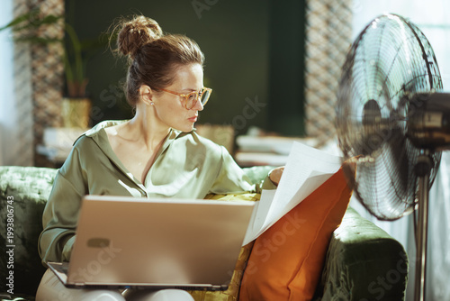 A focused woman with glasses reviews documents while working on her laptop from a green velvet sofa. The running standing fan highlights a productive home office environment in warm weather.