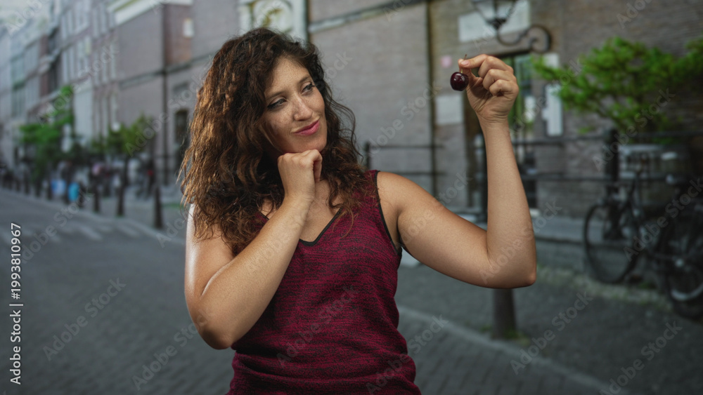 Fototapeta premium Woman holding cherry between fingers with chin on fist on street, raising arm and smiling while wearing burgundy sleeveless top; playful curiosity.