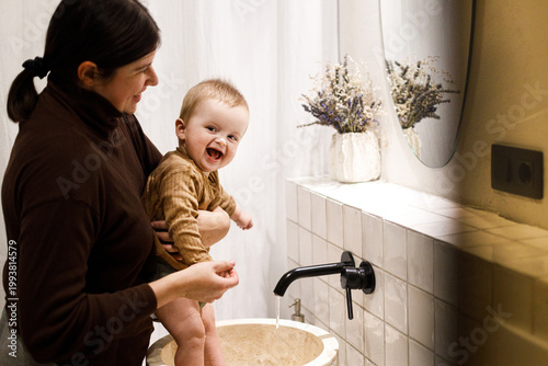 Mother cleaning happy baby infant in modern bathroom after eating complementary food. Family domestic life. Mom washing son cute face with water in sink, joyful moment. Happy Mother's day!