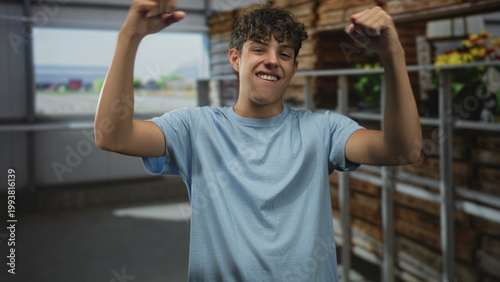 Fotografia Teenage boy flexes biceps and sticks out tongue in a greenhouse building, beside metal shelves and wooden crates with potted flowers; playful confidence