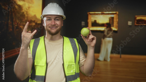 Canvas Print Man engineer in hardhat and high visibility vest holding green apple and making rock horns in building; playful mischief