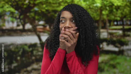 Photography African american woman in long sleeved red shirt gently covers mouth with hands in sunlit forest; surprise
