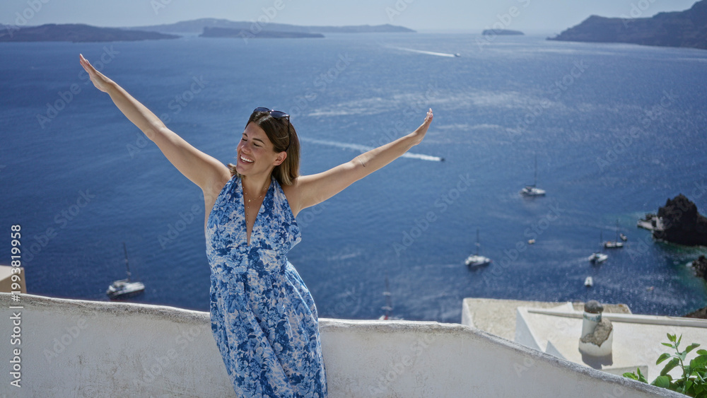 Fototapeta premium Woman smiling with arms out on building terrace overlooking sea and boats in santorini town; vacation relaxation joy.