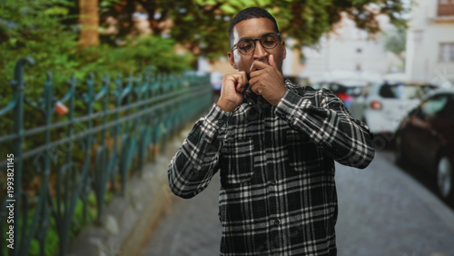 Valokuva Young black man wearing glasses raises arms in celebration after briefly bringing hands to mouth on a street; joy celebration