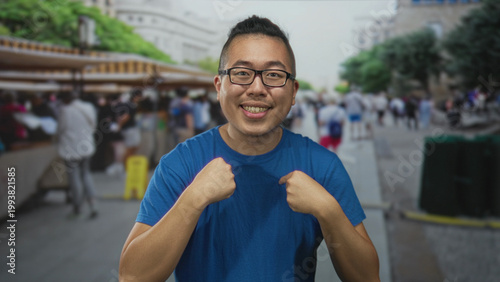 Konstfotografi Man pointing finger to chest at crowded street market, bare forearms visible, wearing blue t shirt and glasses, smiling broadly; confidence pride