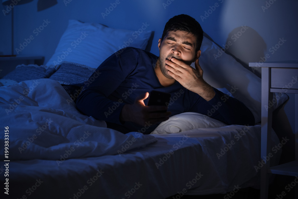 Fototapeta premium Sleepy young man using mobile phone in bed at night