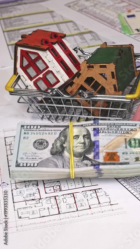 Close-up of house models inside a shopping basket while a stack of US dollars is placed on top, symbolizing real estate purchase, investment decision, and property valuation concept. Vertical video.